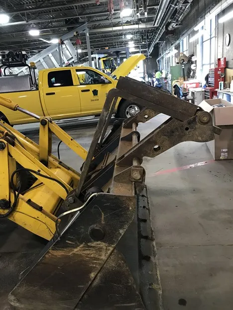 An image showing a side view of a backhoe folding fork attached to a yellow PennDOT construction vehicle.